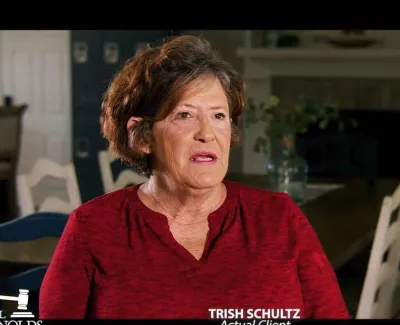 Middle-aged woman in red shirt speaking in a warmly lit dining room with white chairs and dark walls.