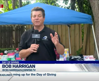 Reporter Bob Harrigan live on air under blue canopy with charity items for Day of Giving event.
