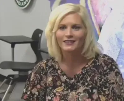 Blonde woman with floral blouse sitting in a classroom with black chairs and a colorful wall decoration.