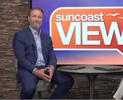 Two hosts seated on chairs in front of a Suncoast View logo with a stone wall background during a TV show.