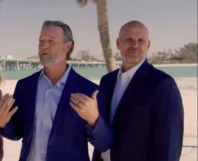 Four professionally dressed people standing on a sunny beach with palm trees and a pier in the background.