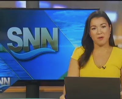 News anchor in a yellow dress presenting live on SNN with a large screen displaying the SNN logo behind her.