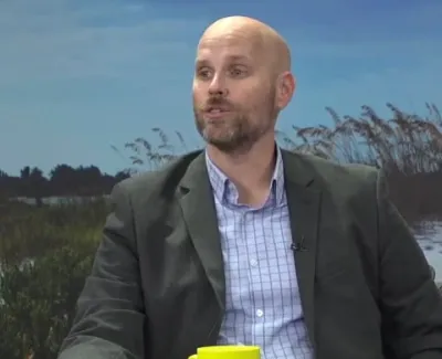 Bald man in a suit jacket speaking with a beach and sea grass backdrop during an interview.