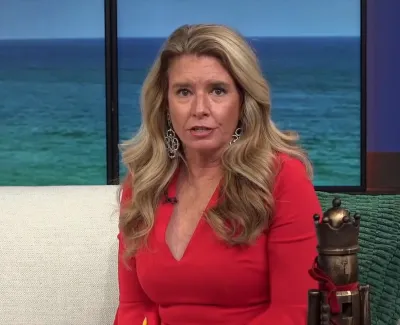 Woman in a red dress with long blonde hair sitting on a white couch with ocean view behind her.