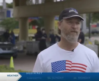 Man wearing an American flag shirt and cap being interviewed outdoors near a bridge on a sunny day.