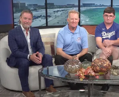 Four TV show hosts sitting on a couch with a glass coffee table and ocean view background in a studio setting.