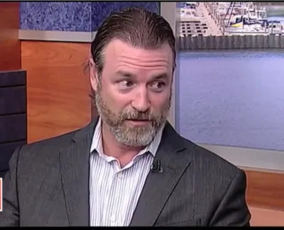 Man in gray suit and striped shirt speaking on Suncoast View TV show with waterfront background.