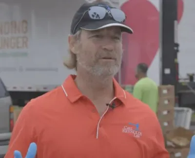 Man in orange shirt and cap speaking outdoors near trucks with Ending Hunger logo in background