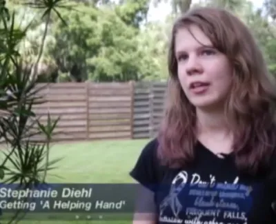 Young woman speaking outdoors with greenery and wooden fence in background, wearing black graphic t-shirt.