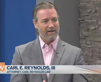 Man in grey suit and pink shirt speaking on a TV news set with stone wall background and lower third text