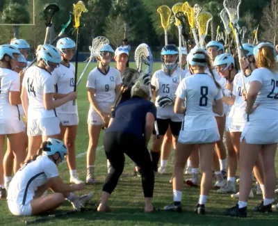 Women lacrosse team in white uniforms gathers around coach on grass field during practice
