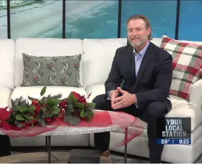Two people sitting on a white couch with festive holiday decorations and pillows in a TV studio.