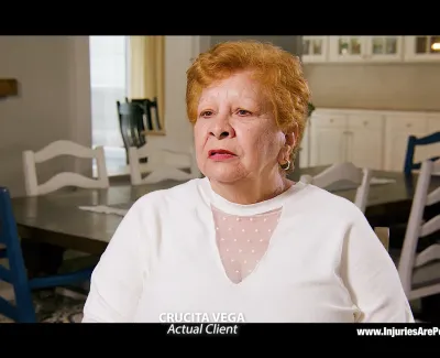 Elderly woman with short red hair in white sweater speaking in a dining room setting for a law firm testimonial.