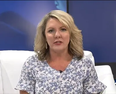 Woman news anchor with blonde hair wearing a floral blouse seated on a white couch with blue background
