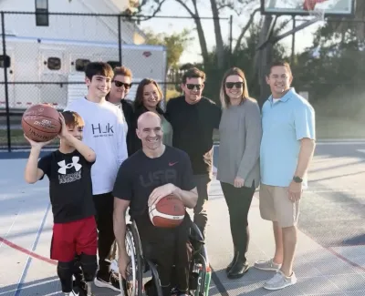 A diverse group of smiling people, including a man in a wheelchair holding a basketball, on an outdoor basketball court.