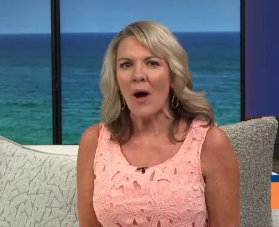 Female TV host in pink lace dress speaking on a coastal-themed set with ocean view background