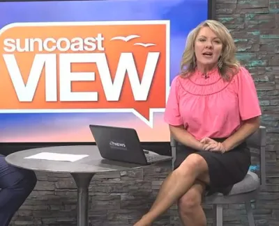 Two hosts sitting and talking on the Suncoast View TV show set with laptop and large logo backdrop.