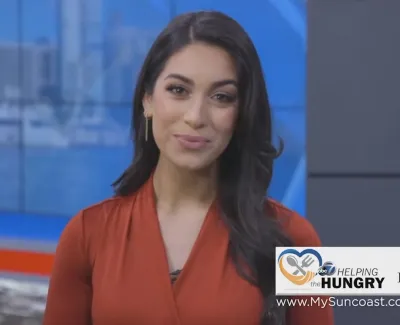 Female news anchor in orange blouse presents from studio with harbor view and HELPING the HUNGRY charity logo.