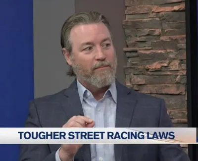 Man in a suit discussing tougher street racing laws on a news segment with informational banner and QR code.