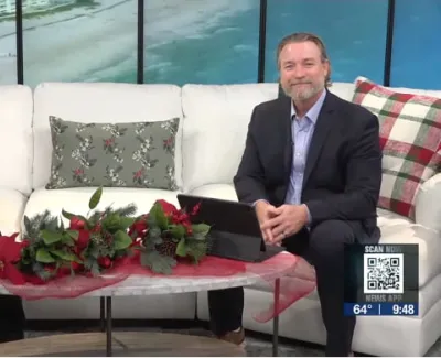 Two hosts sit on a white couch with holiday decorations on the coffee table in a TV studio with a beach backdrop.