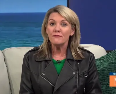 Blonde woman in black leather jacket sitting on a couch with ocean backdrop during a TV segment.