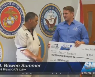 Man presenting a $1,000 donation check to another man in front of military branch emblems and TV station logo.