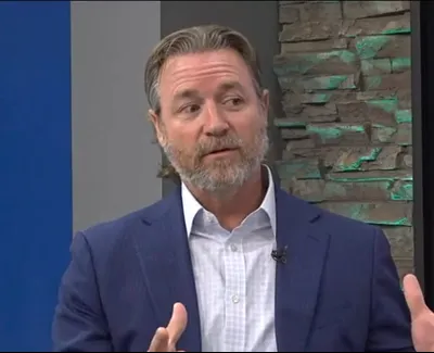 Bearded man in blue suit jacket discussing on a news broadcast set with digital display and stone wall background