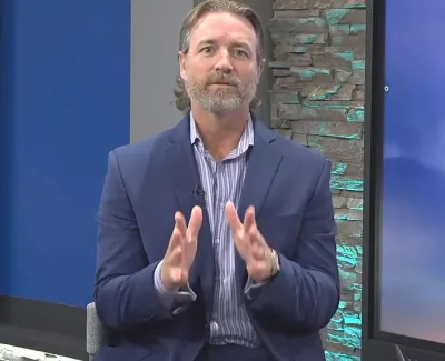 Man in blue blazer and striped shirt speaking on a TV news set with a sunny view graphic and local station info