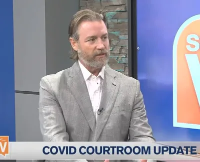 Man in gray suit giving COVID courtroom update on Suncoast View TV show set with stone wall background.