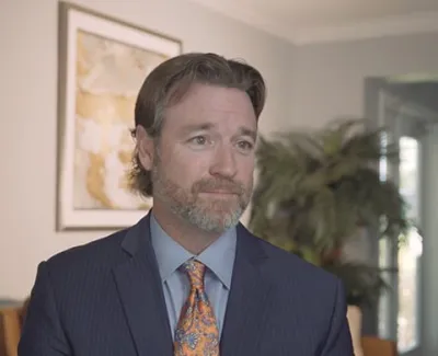 Man with beard and mustache wearing a suit and orange patterned tie in a professional office setting.
