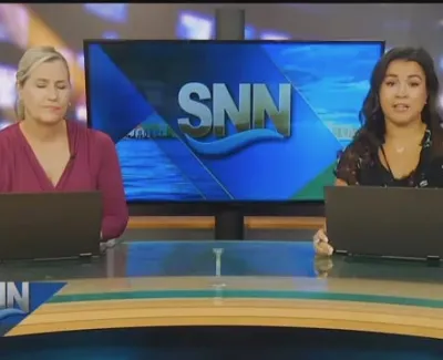 Two news anchors at a news desk with laptops, SNN logo on screen, time and temperature displayed in corner.
