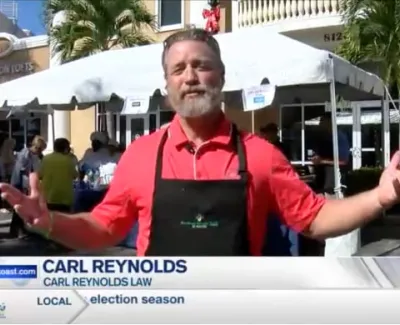 Man in red shirt and black apron speaking at outdoor event with tents and palm trees in background