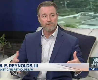 Attorney Carl E. Reynolds III in a navy suit speaking during a local TV interview with logo and lower-thirds displayed.