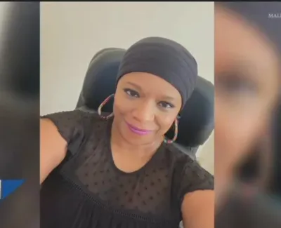 Smiling woman wearing black headwrap and top sitting in chair with hoop earrings and pink lipstick.