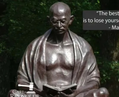 Bronze statue of Mahatma Gandhi reading a book outdoors with a quote about service and self-discovery.