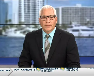 Male news anchor in suit and tie presenting weather forecast with city waterfront background on screen.