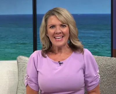 Smiling blonde woman in a lavender blouse sits on a couch with ocean view in the background.