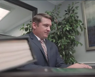 Professional man in suit working at office desk with large book, office plant, and framed artwork in background.