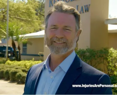 Smiling man in suit standing outside Carl Reynolds Law office with greenery and parking lot in background