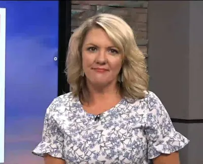 Blonde woman wearing floral blouse standing in front of a news studio background with orange and blue colors
