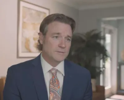 Man in a blue suit and colorful tie speaking in a well-lit modern office with wall art and plants.
