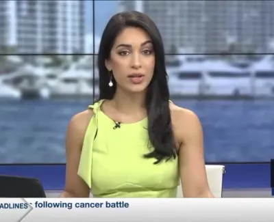 Female news anchor in green dress delivering live broadcast on local TV news studio with city waterfront background.