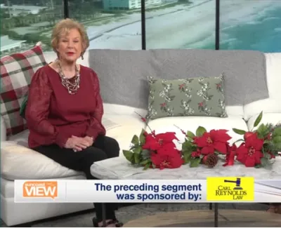 Two hosts seated on a white couch in a TV studio with festive poinsettia decorations and a beach view background.