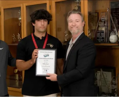 Student holding award plaque for Student Athlete of the Month, flanked by two men in front of trophy case.