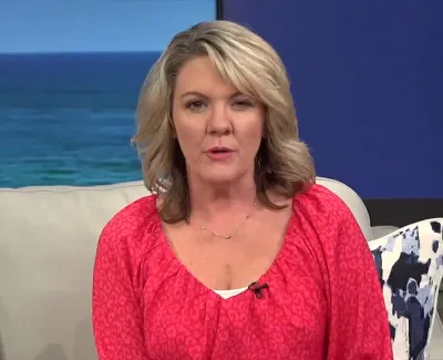 Blonde woman in a bright pink blouse sitting on a beige couch with ocean background and plant nearby