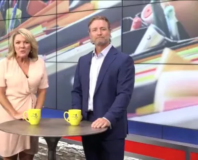 Two news anchors standing by a round table with yellow mugs in a modern studio with a stone wall and large screen.