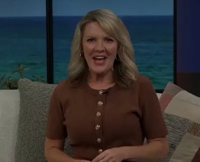 Blonde woman in brown top sitting on beige sofa with cushions and ocean view in background.