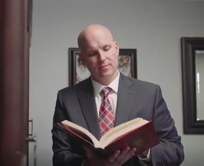 Man in business suit reading a large hardcover book in a modern office setting with framed artwork.