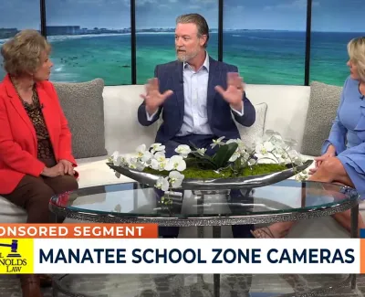 Three news anchors discussing Manatee school zone cameras on a TV set with ocean view background.