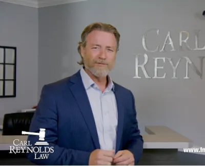 Male lawyer in blue suit standing in front of Carl Reynolds Law office logo with legal gavel symbol.